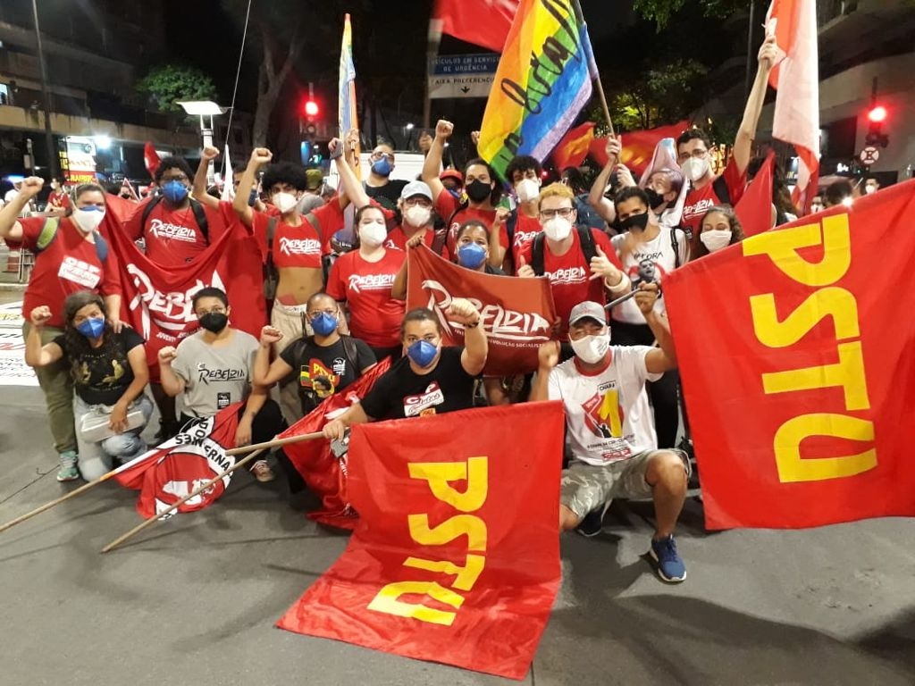 Jovens agrupados segurando bandeiras do PSTU, do coletivo Rebeldia e do movimento LGBTQIA +.  A maioria está com blusas que estampam a logo do coletivo e todos eles estão de máscaras. Muitos estão com a mão levantada em sinal de resistência.