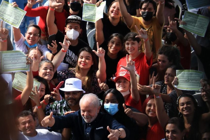 Aglomeração de jovens, muitos sem máscara, em ambiente aberto, fazendo um “L” com as mãos e segurando Títulos de Eleitores. O ex-presidente Lula aparece na parte inferior da fotografia, no meio das pessoas.