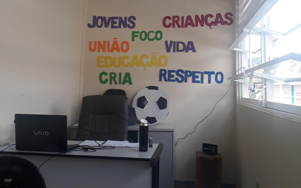 Uma sala, que tem uma mesa e uma cadeira vazia. Na parede atrás está escrito “jovens”, “crianças”, “foco”, “união”, “vida”, “educação”, “CRIA”, “respeito” e tem a ilustração de uma bola de futebol.